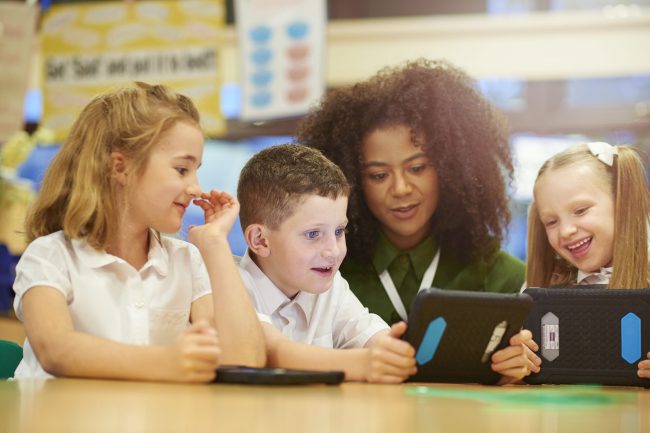 three primary aged schoolchildren sit at their desks and look engrossed in the work that they are doing on their digital tablets. Their schoolteacher is looking over their shoulder and joining in with pupils . They are all wearing white school shirts
